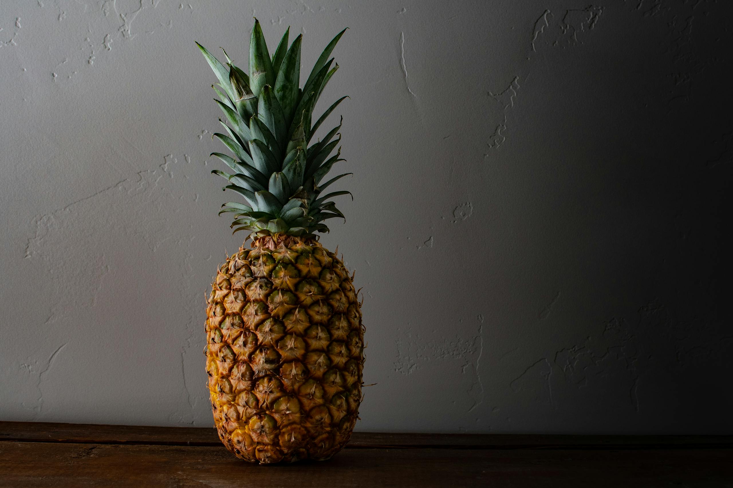 A ripe pineapple placed on a wooden surface against a textured wall, capturing its natural, fresh appeal.